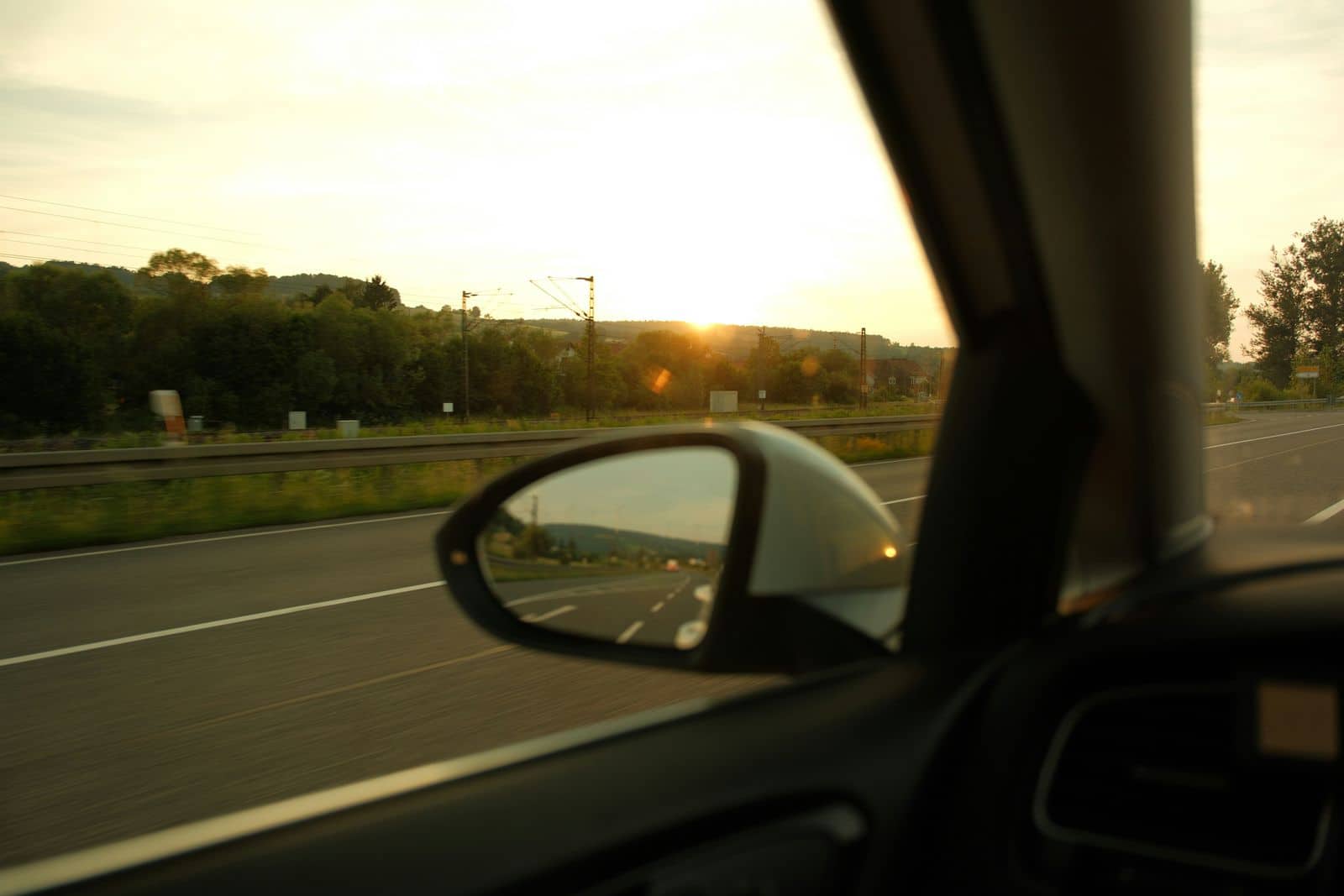 A highway at sunset seen from the driver's seat, with the side mirror catching the light