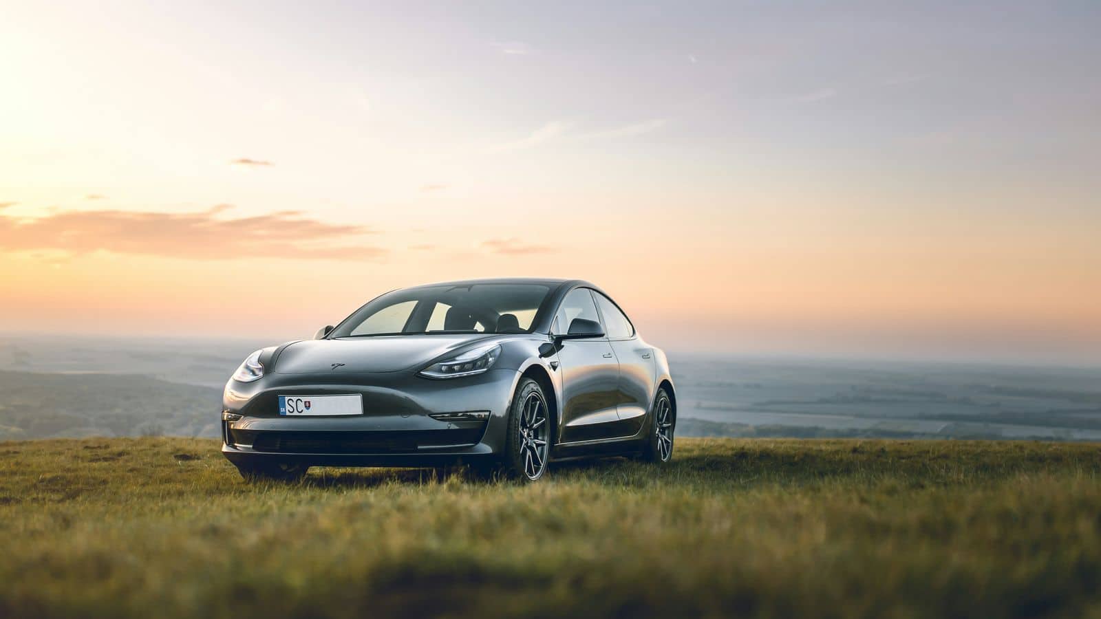 An electric car parked on grass at dusk, with hills and an orange sky behind it